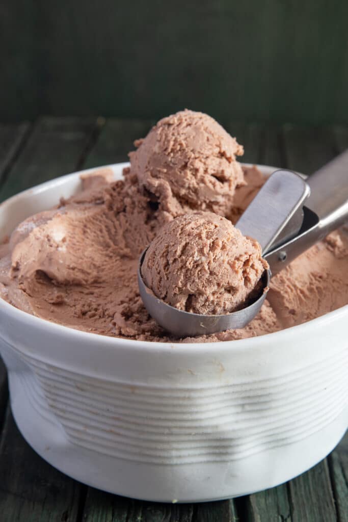 No churn chocolate ice cream in a white bowl with an ice cream scoop containing an ice cream ball.