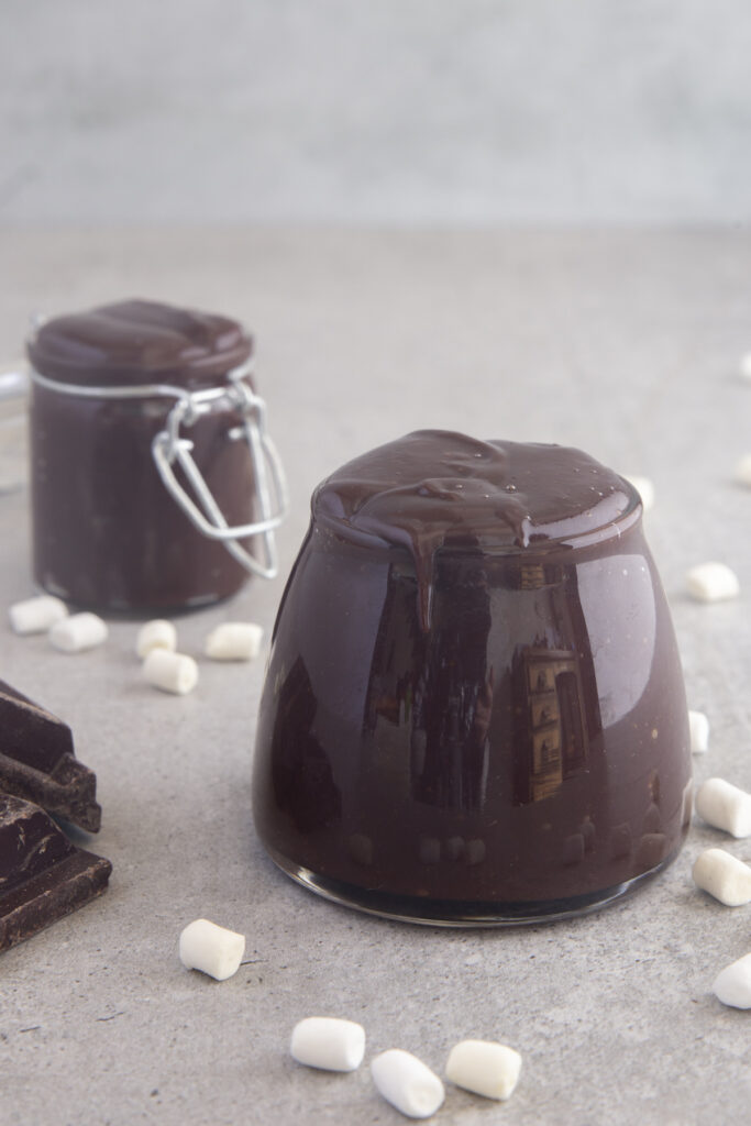 Fudge sauce in a glass jar with another jar of sauce behind it.
