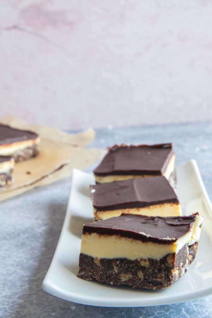 Three nanaimo bars on a white plate with the rest of the bars on parchment paper in the back.