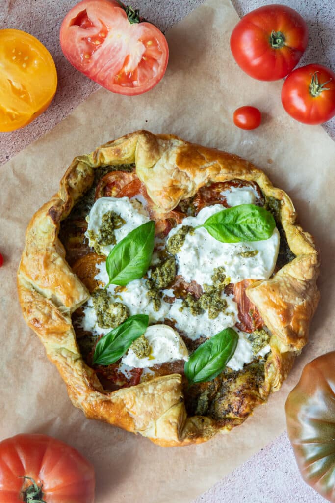A whole tomato galette on brown parchment paper and heirloom tomatoes around it.