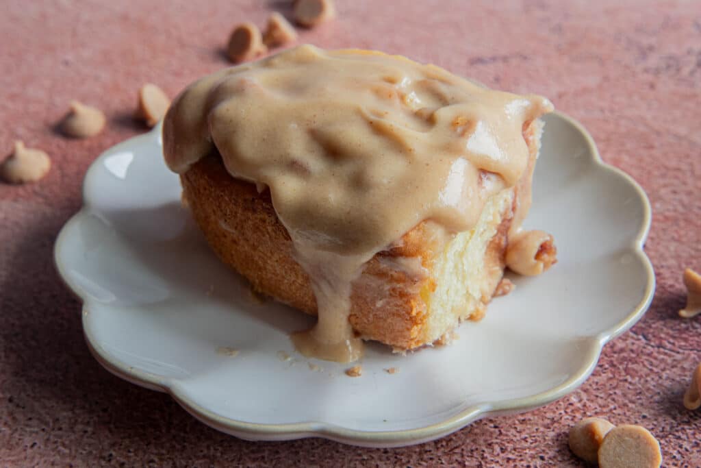 A peanut butter cinnamon roll with glaze on a white plate and peanut butter chips around it.