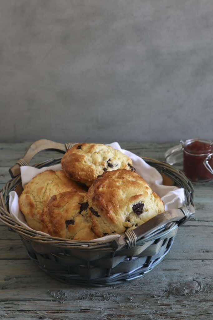 Four chocolate chip scones in a blue basket with jam in a glass on the right side.