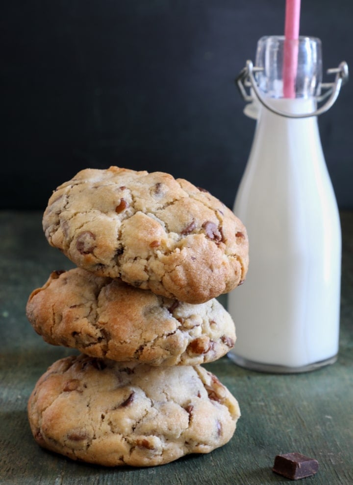 Three chocolate chip cookies stacked and a glass bottle of milk with a pink straw behind them.