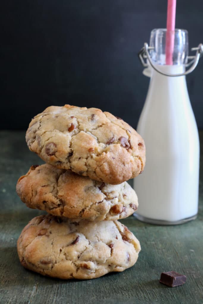 Three chocolate chip chunky cookies one on top of the other with a glass bottle of milk behind them.