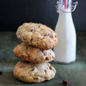 Three chocolate chip chunky cookies stacked and a bottle of milk in the back.