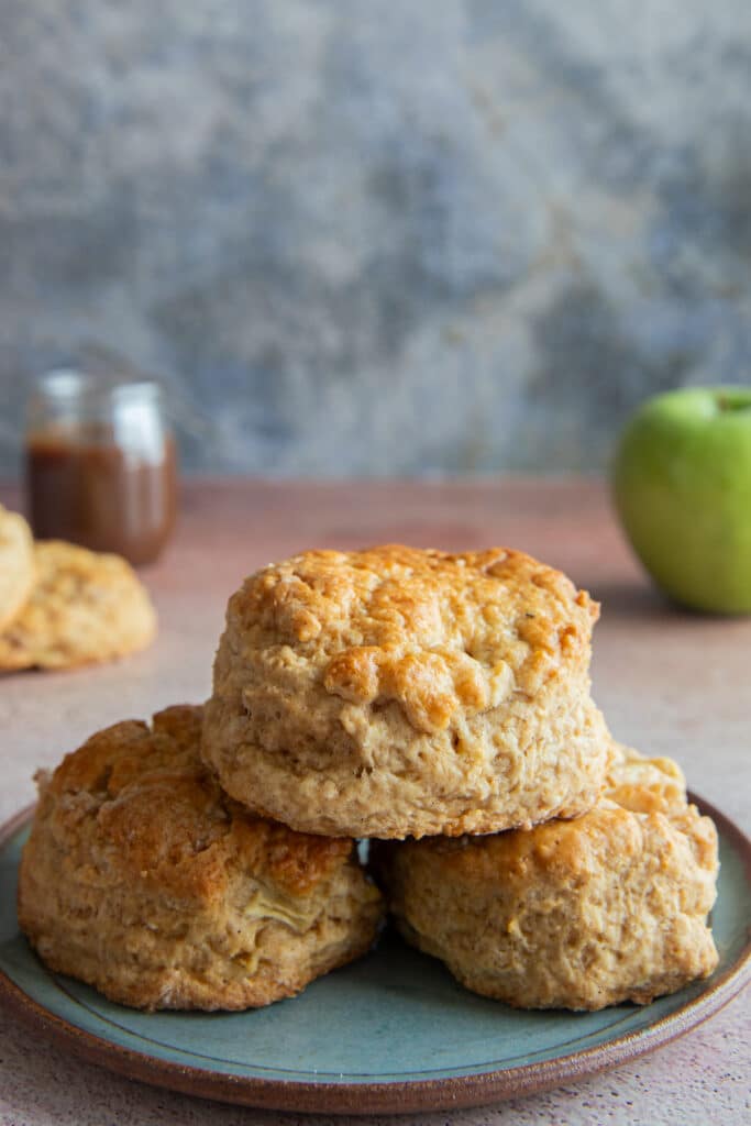 Three scones on a green plate with more scones and a jar of caramel sauce on the top left side and a green apple on the top right side.