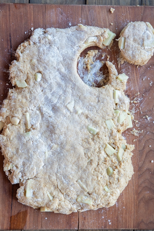 Apple and caramel scone dough flatten on a wooden board with one circle cut into it.