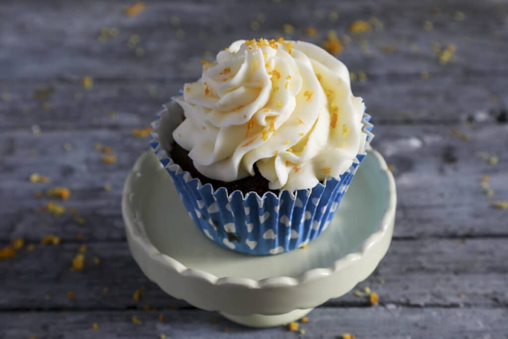 A chocolate orange cupcake in a blue cupcake liner on a white cake stand.