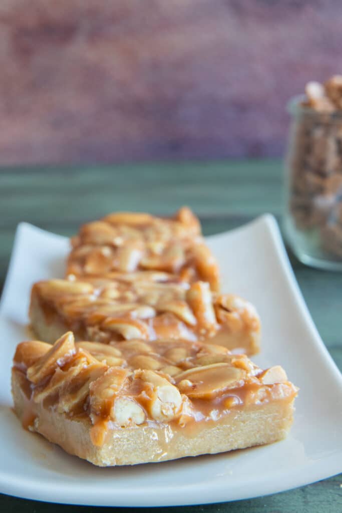 Three peanut squares on a white plate with a jar full of butterscotch chips in the back.
