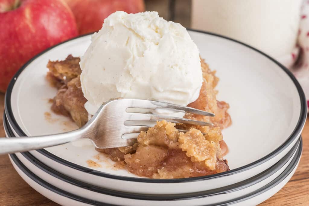 A fork cutting into a slice of bottomless apple pie with a scoop of ice cream on a stack of white plates.