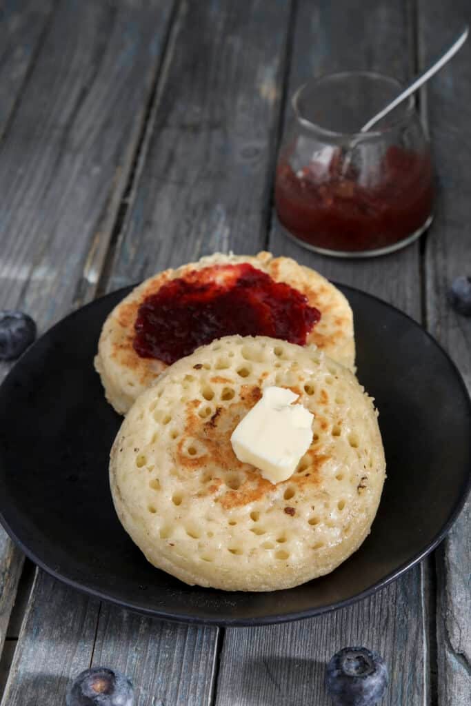 Two crumpets on a black plate with butter on one and jam on the other one.