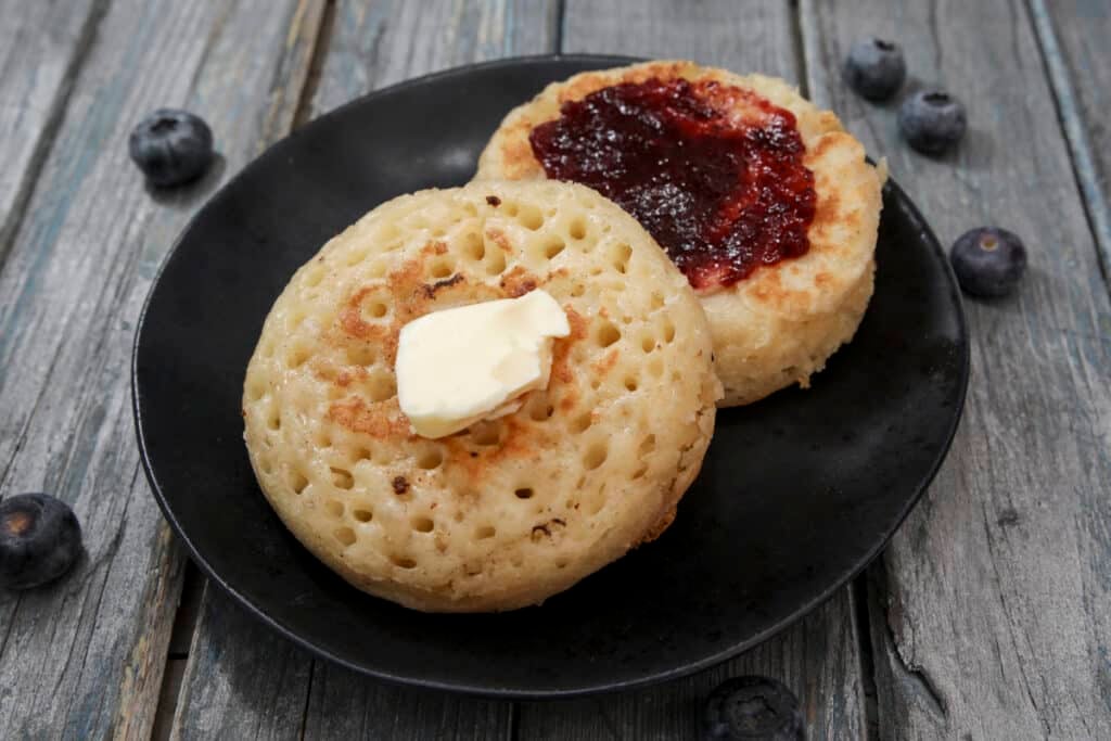 Two crumpets on a black plate with blueberries scattered around.