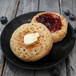 Two crumpets with butter and jam on a black plate.