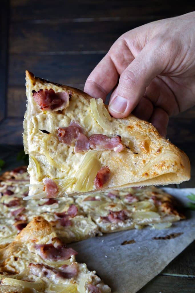 A hand holding a slice of flammkuchen with the rest of it in the back on a sheet of parchment paper.