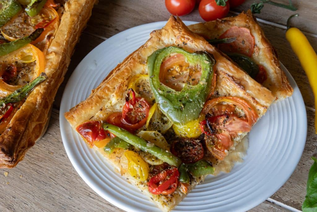 A slice of tomato and mascarpone tart on a white plate with the rest of the tart on the upper left side.